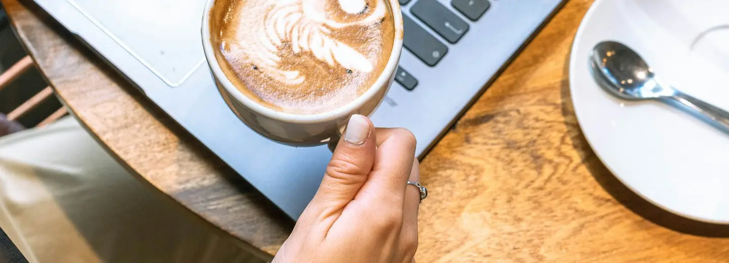 Hand holding a latte over a laptop on a café table