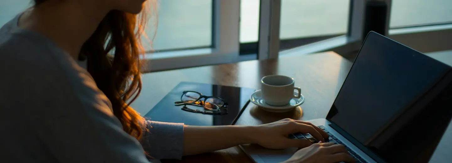 Person working on a laptop by a window with a coffee