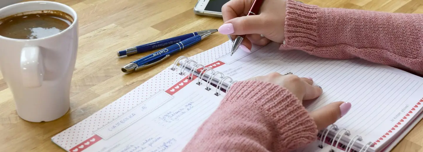 Hands filling in a weekly planner with coffee on the table