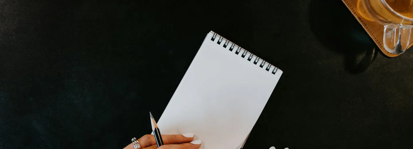 Hands with a pen over a blank notepad next to a drink